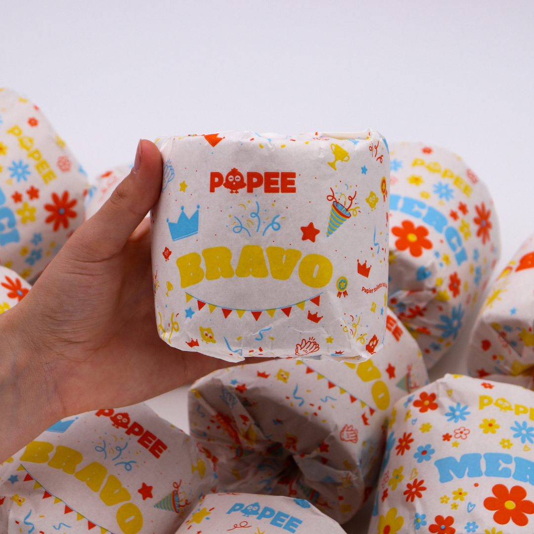 Main tenant un rouleau de papier toilette Popee ‘BRAVO’ sur fond blanc, détails d’un emballage festif et coloré.