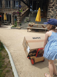 Enfant tirant un chariot avec un carton Popee devant un gîte en pierre.
