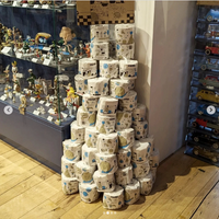 Une pyramide de rouleaux de papier toilette Popee illustrés en bande dessinée, empilée au sol dans une librairie, à côté d'une vitrine de figurines de collection.