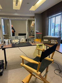 Loge de maquillage en coulisses avec miroir triptyque, produits cosmétiques et boîte de mouchoirs Popee sur la table, chaise de réalisateur au premier plan et malle de rangement.