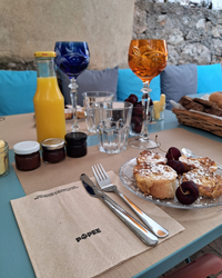 Une table de petit-déjeuner dressée avec une serviette en papier Popee, des toasts briochés aux cerises, un jus d'orange, des confitures et des verres en cristal colorés, sur fond de mur en pierre et coussins bleus.