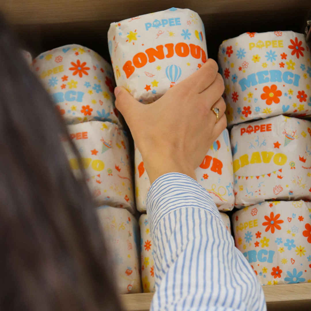 Une personne attrape un rouleau Popee ‘BONJOUR’ dans une étagère pleine de papiers toilette colorés et festifs.