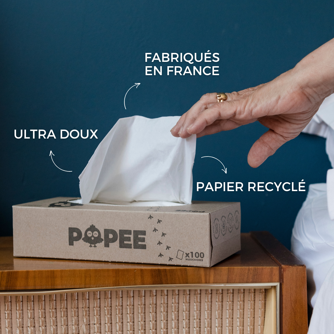 Main prenant un mouchoir recyclé Popee, boîte en carton sur table bois, fond bleu.