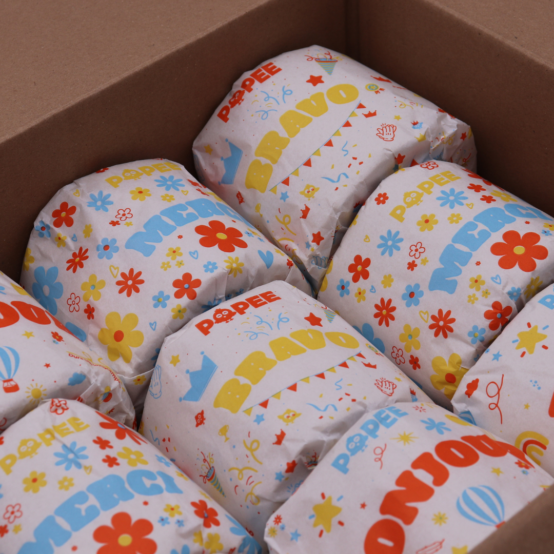 Rouleaux de papier toilette Popee aux messages colorés ‘BRAVO’, ‘MERCI’ et ‘BONJOUR’, soigneusement rangés dans un carton.
