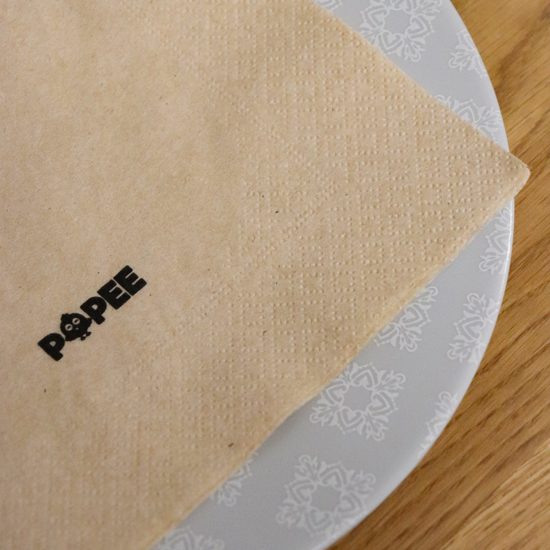Serviette de table recyclée Popee posée sur une assiette, texture naturelle et logo imprimé.