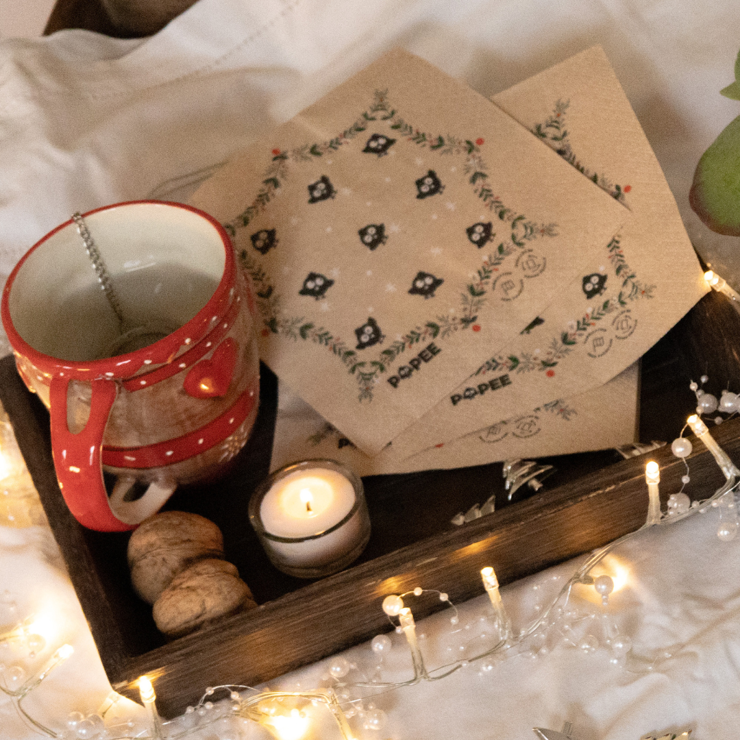Serviettes en papier recyclé POPEE à motif de Noël, posées sur un plateau avec tasse et bougie dans une ambiance chaleureuse.